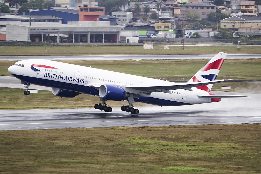Um avião inicia a decolagem em uma pista de decolagem. O avião é branco e possui detalhes em azul e vermelho e, na sua lateral, a logo da British Airways.